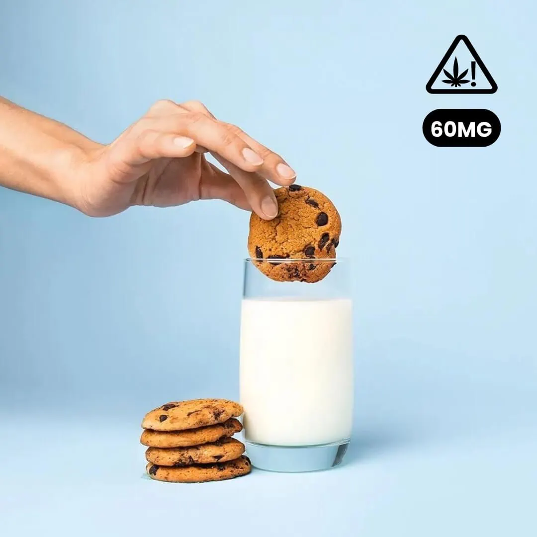 A person is putting a Baked Delta-9 Chocolate Trip cookie into a glass of milk.