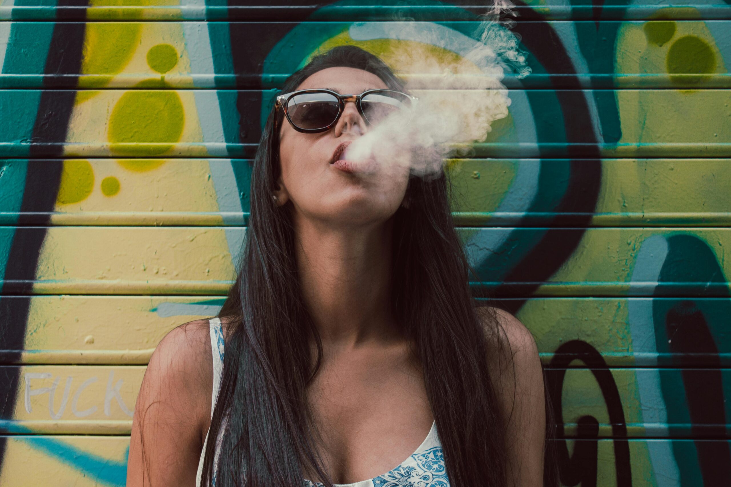 Woman exhaling smoke in front of a graffiti-covered wall.