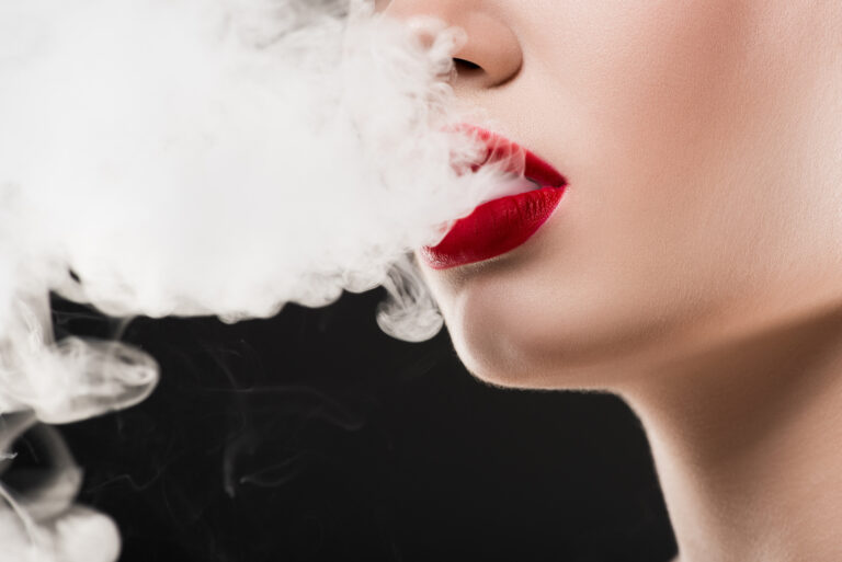 Close-up of a woman with red lipstick exhaling smoke against a black background, questioning "Is HHC safe?