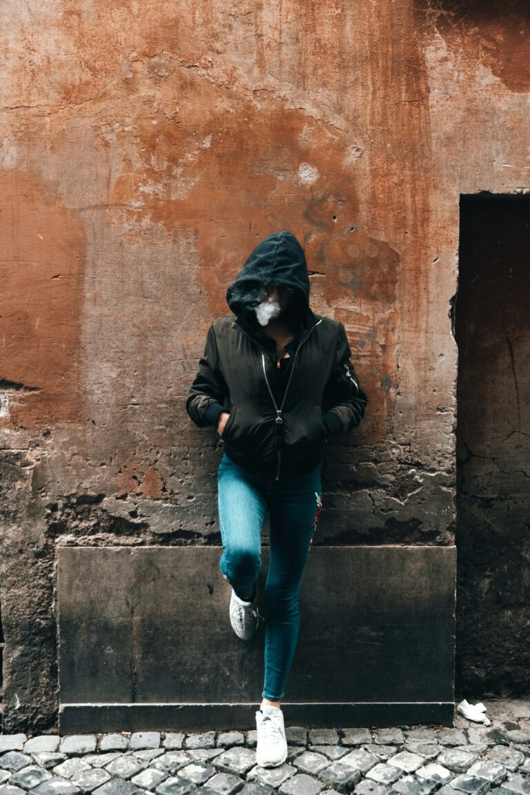 A person in a hooded jacket and jeans stands against a textured orange wall, with one foot propped on the wall, face obscured by shadow. Will HHC get you higher than Delta-8